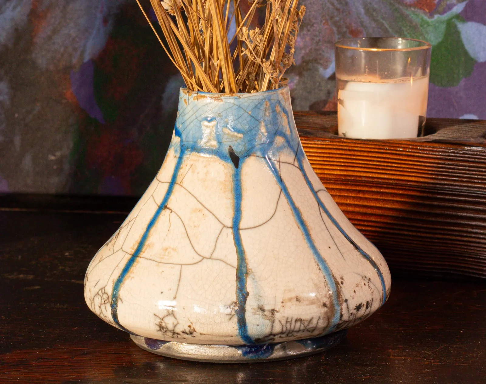 A squat onion-shaped vase in white crackle glaze with cobalt blue running down from the rim, holding dried lavender, beside a lit candle.