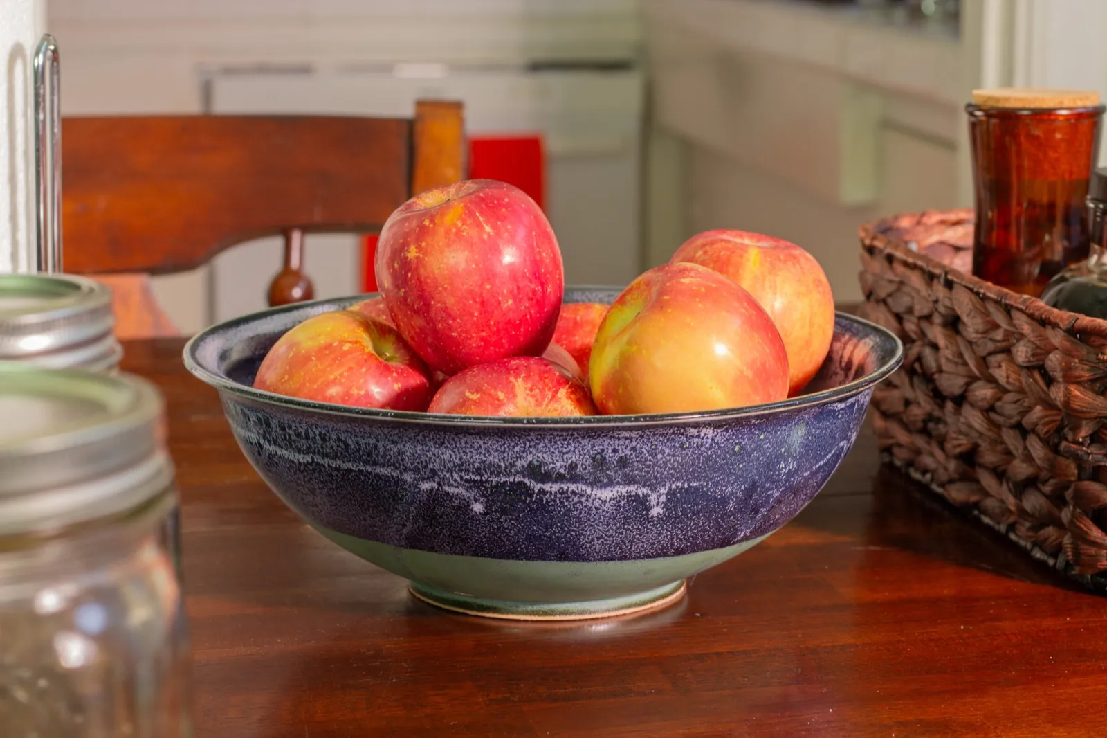 A wide stoneware bowl glazed deep aubergine over celadon, holding six red and yellow apples, on a dark wooden table between mason jars and a basket.