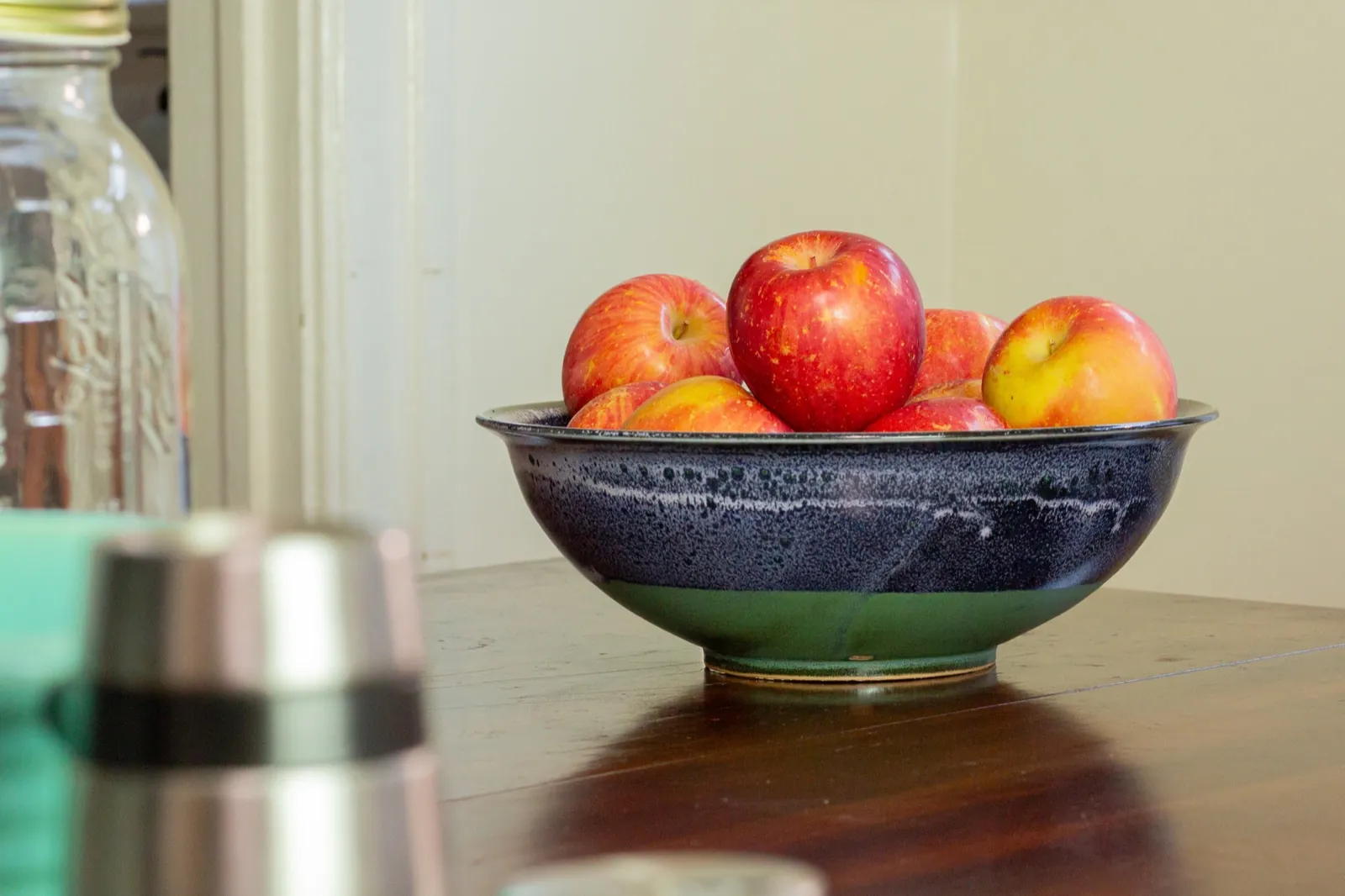 The bowl of apples seen across the table, a glass jar soft in the foreground.