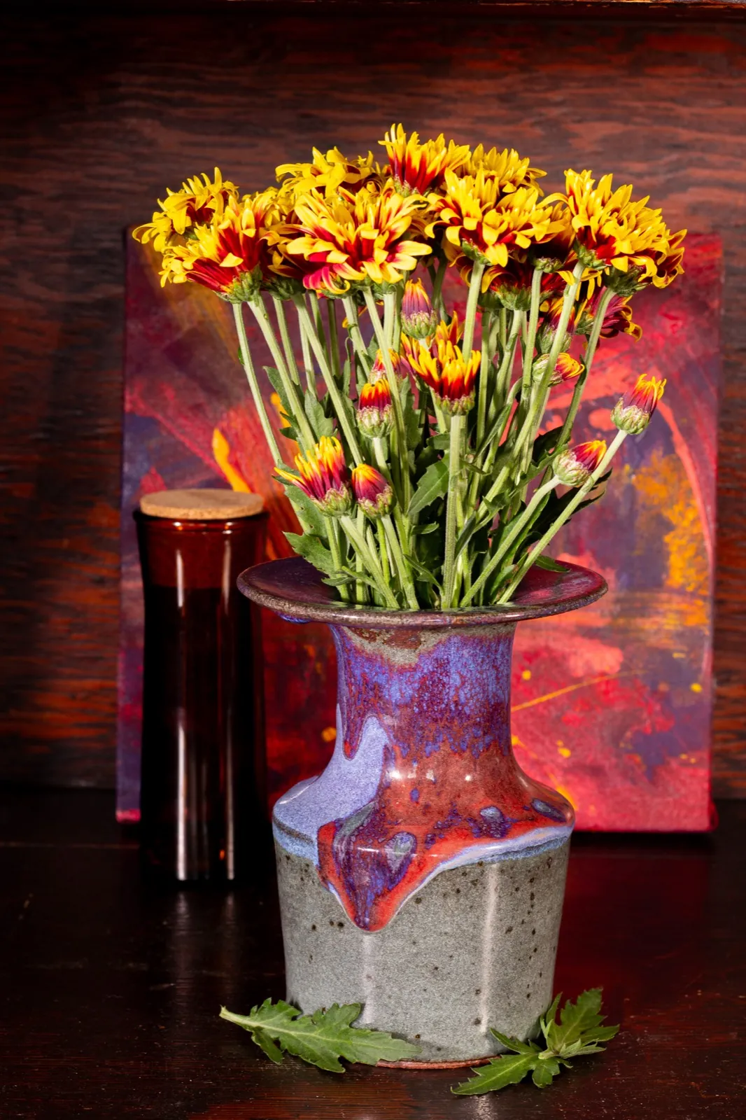 The flanged bottle holding yellow and red chrysanthemums, against a red and purple painting.